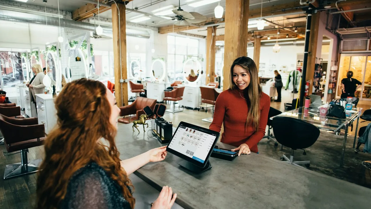 A customer stands at a shop counter while a staff member beside a tablet payment terminal serves her in a bright, open salon.