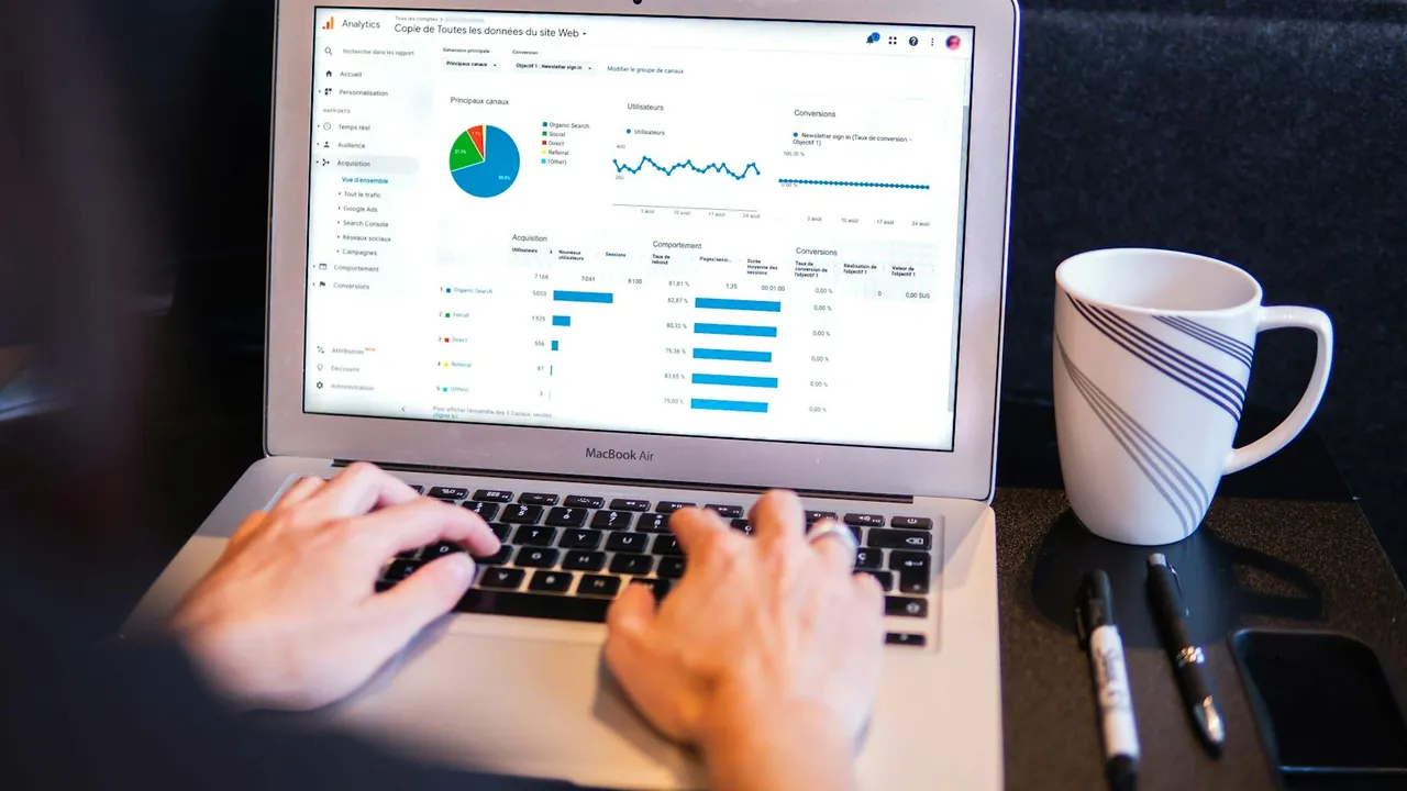 Hands typing on a laptop that shows a dashboard with charts and tables, next to a coffee mug and pens.