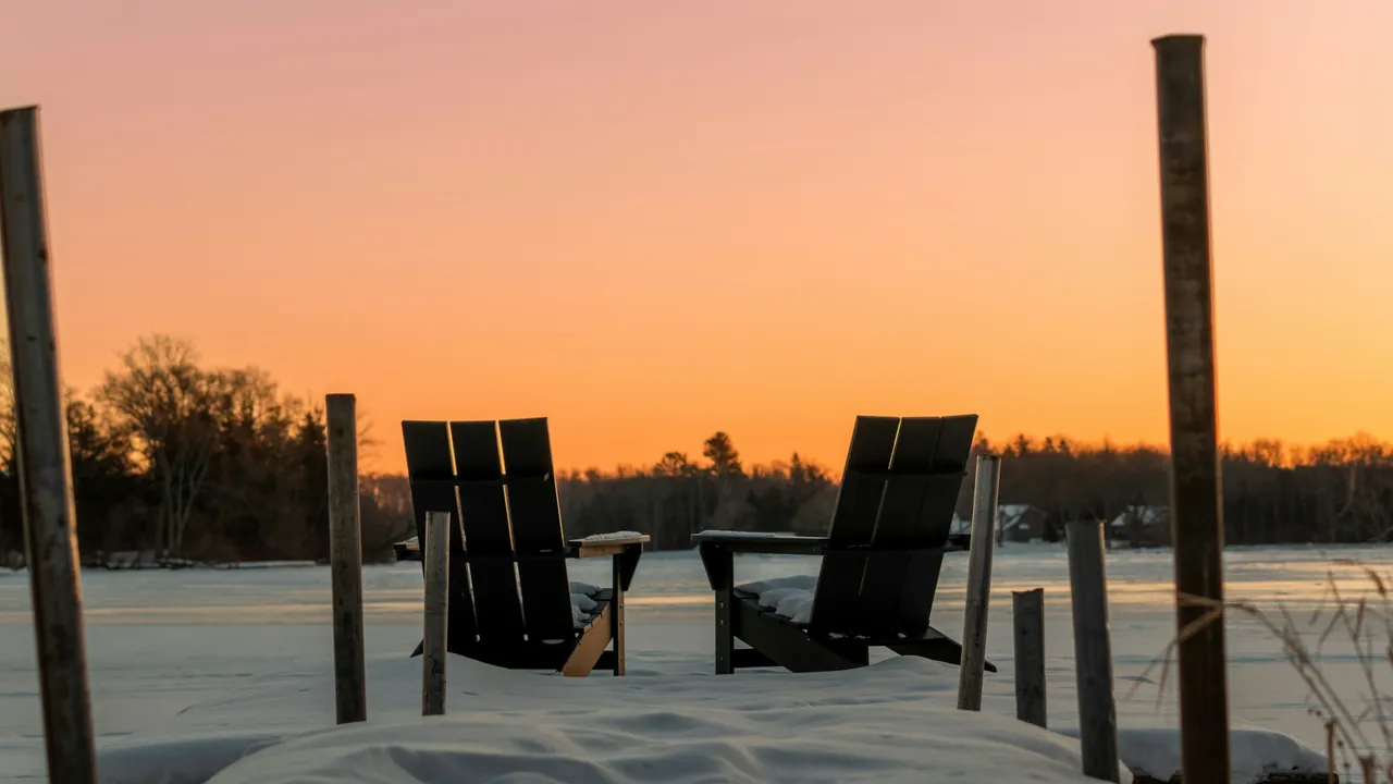 Two Muskoka chairs on a snow-covered dock at sunset, looking over a frozen lake.