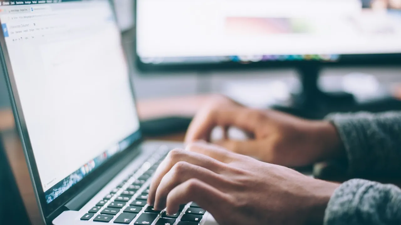 Close view of a person's hands typing on a laptop keyboard at a desk, with a second screen behind.