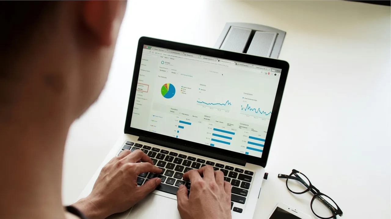 Person typing on a laptop that shows charts on a reporting dashboard, with glasses and a phone beside it.