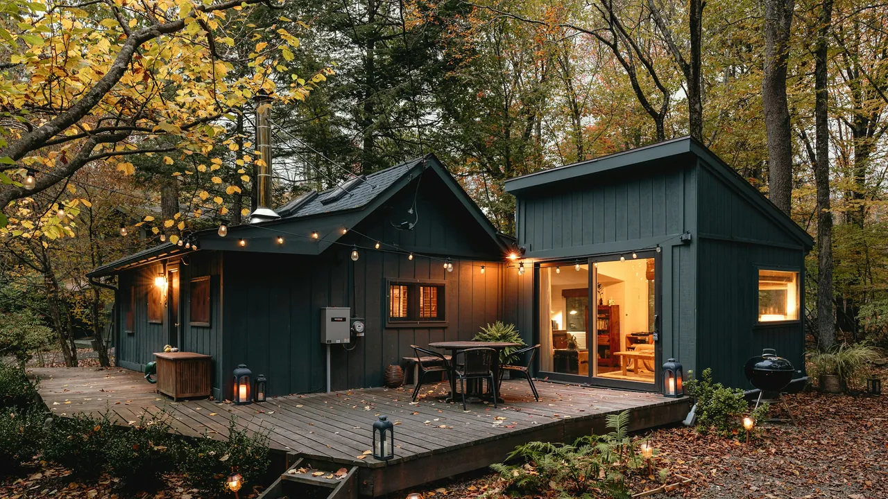 Two dark green cabins with a lit deck and outdoor dining table, surrounded by autumn forest at dusk