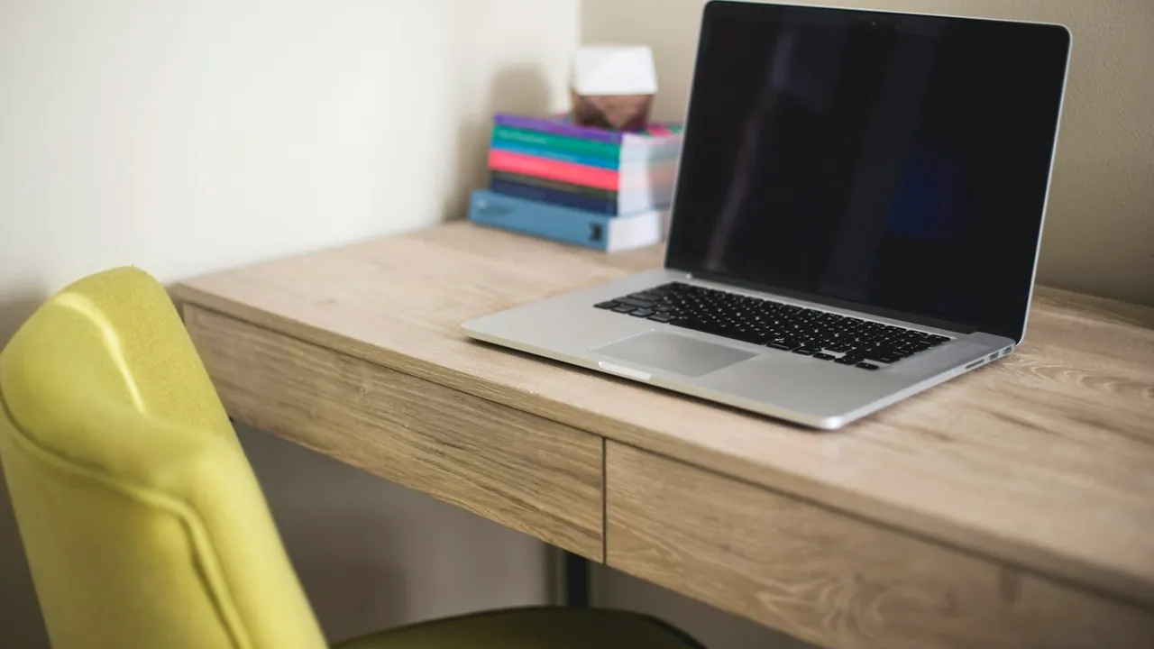 A silver laptop open on a light wooden desk beside a stack of books and a yellow chair.