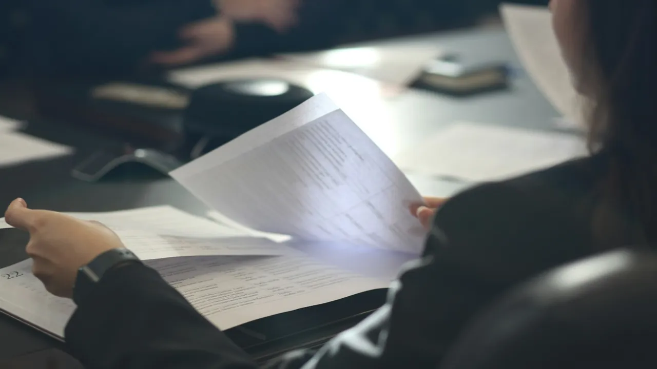 A person seated at a desk holds up and reads a printed document with several pages on the table.
