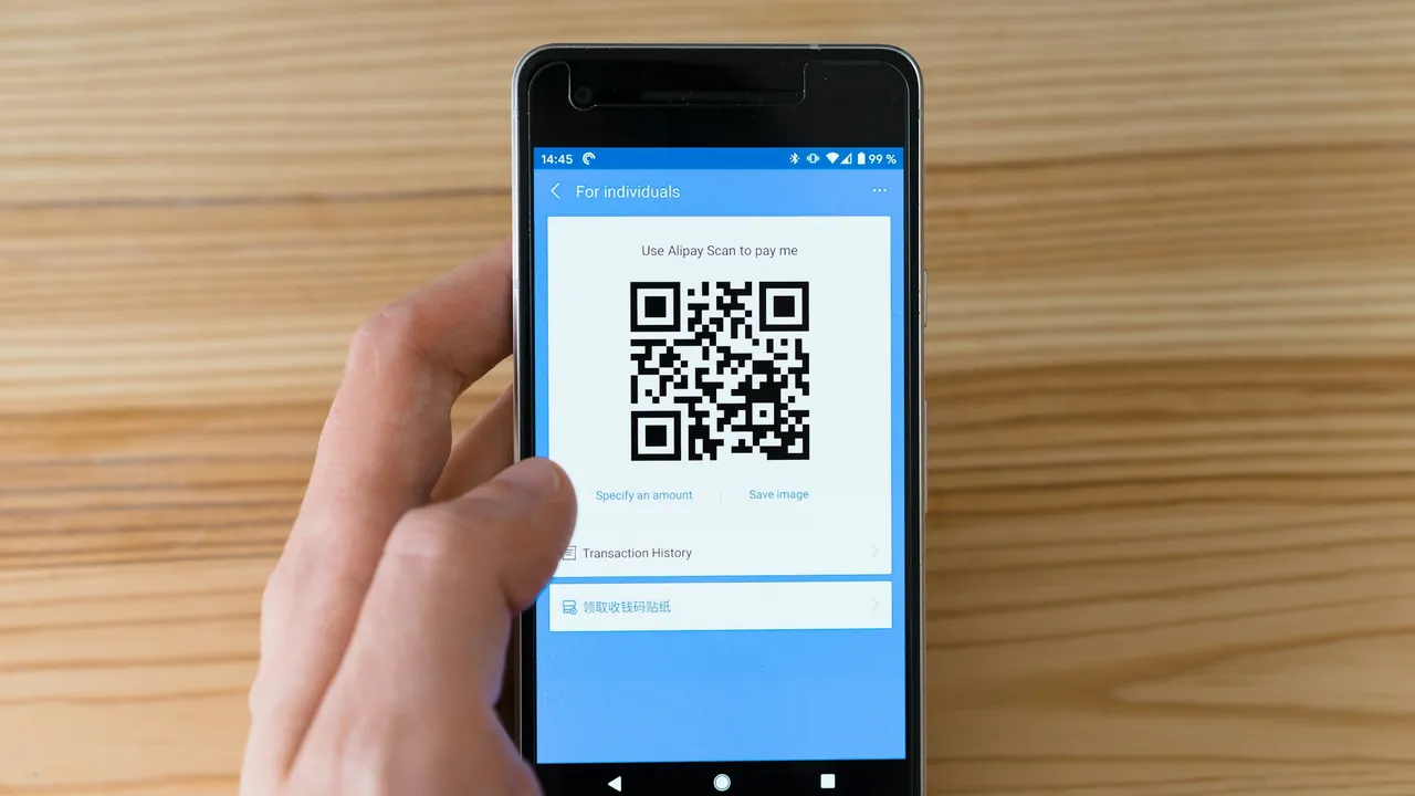 A hand holding a phone that shows a QR code, over a wooden table.