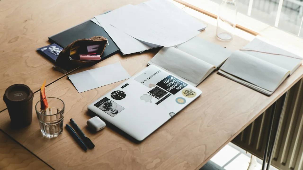Loose papers, notebooks and a closed laptop spread across a wooden desk beside a window, with a coffee cup and pens.