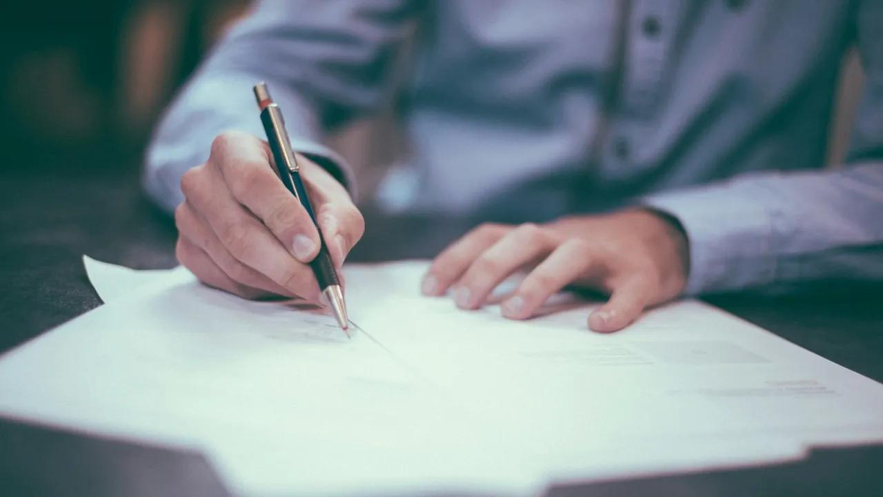 A person in a blue shirt signs a paper document on a desk with a pen.