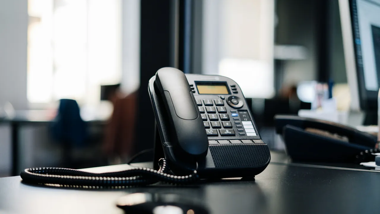 A black corded desk telephone with a keypad sits on a dark desk in a bright office.